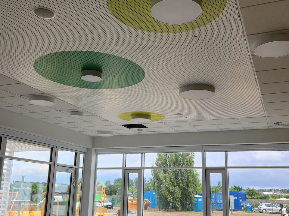 Sala przedszkolna z perforowanym sufitem akustycznym w zielone koła, okrągłymi lampami i nawiewnikiem wirowym, za oknami plac zabaw