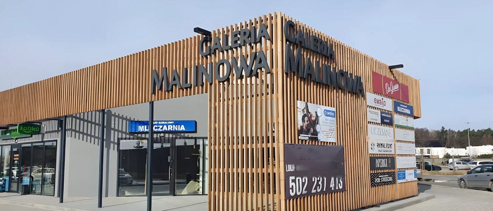 Pawilon handlowy Galeria Malinowa w Dąbrówce z elewacją z drewnianych lameli i szyldami najemców